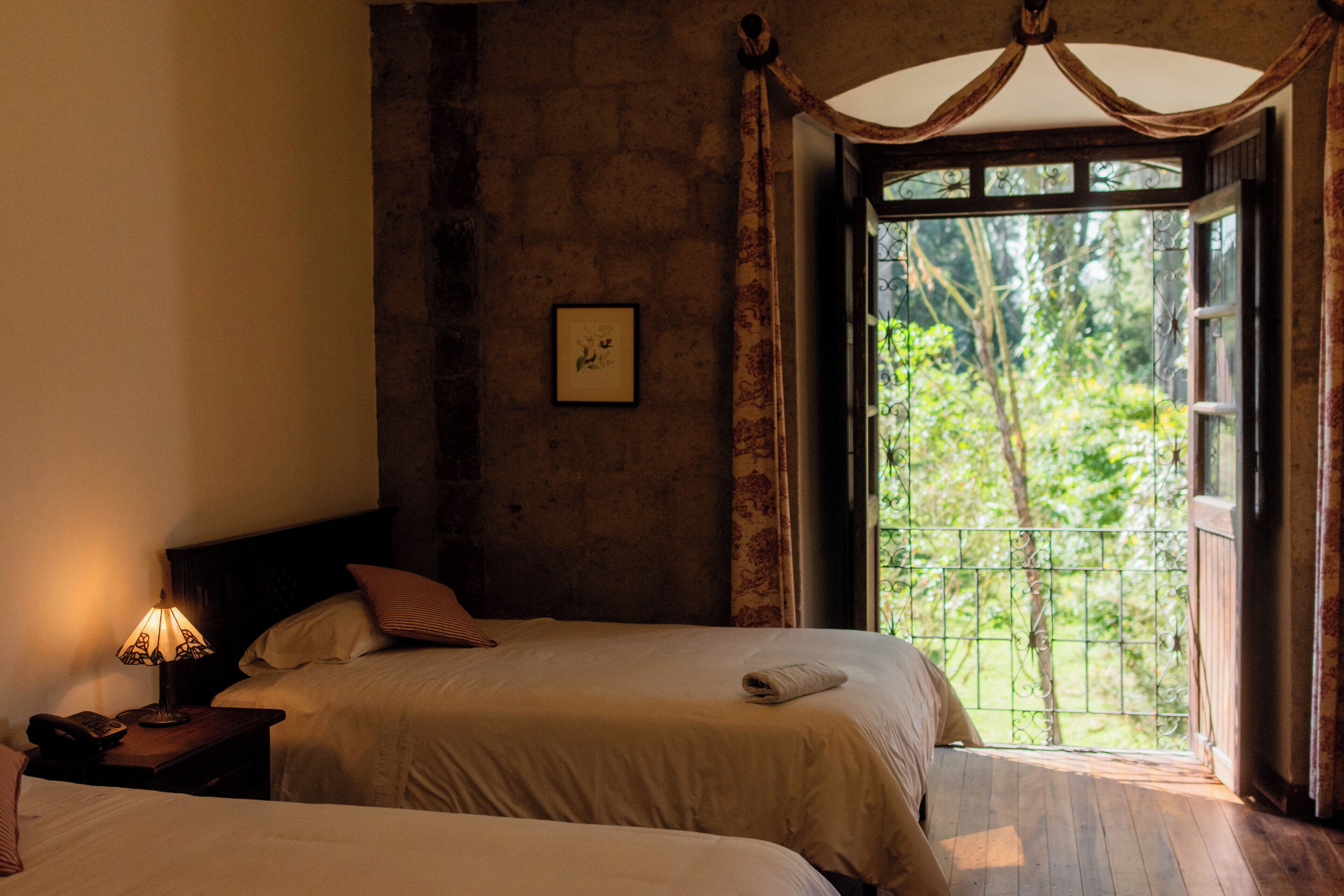 Quarto com duas camas, uma mesa de cabeceira com abajur e uma janela com vista para o verde.