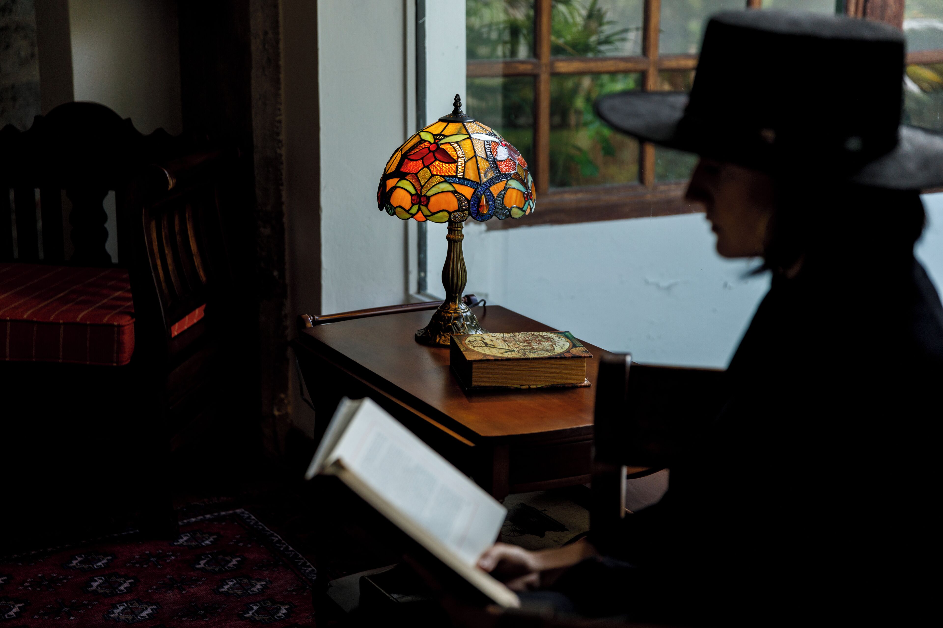A person reading a book at a wooden table with a stained glass lamp and a book beside it.