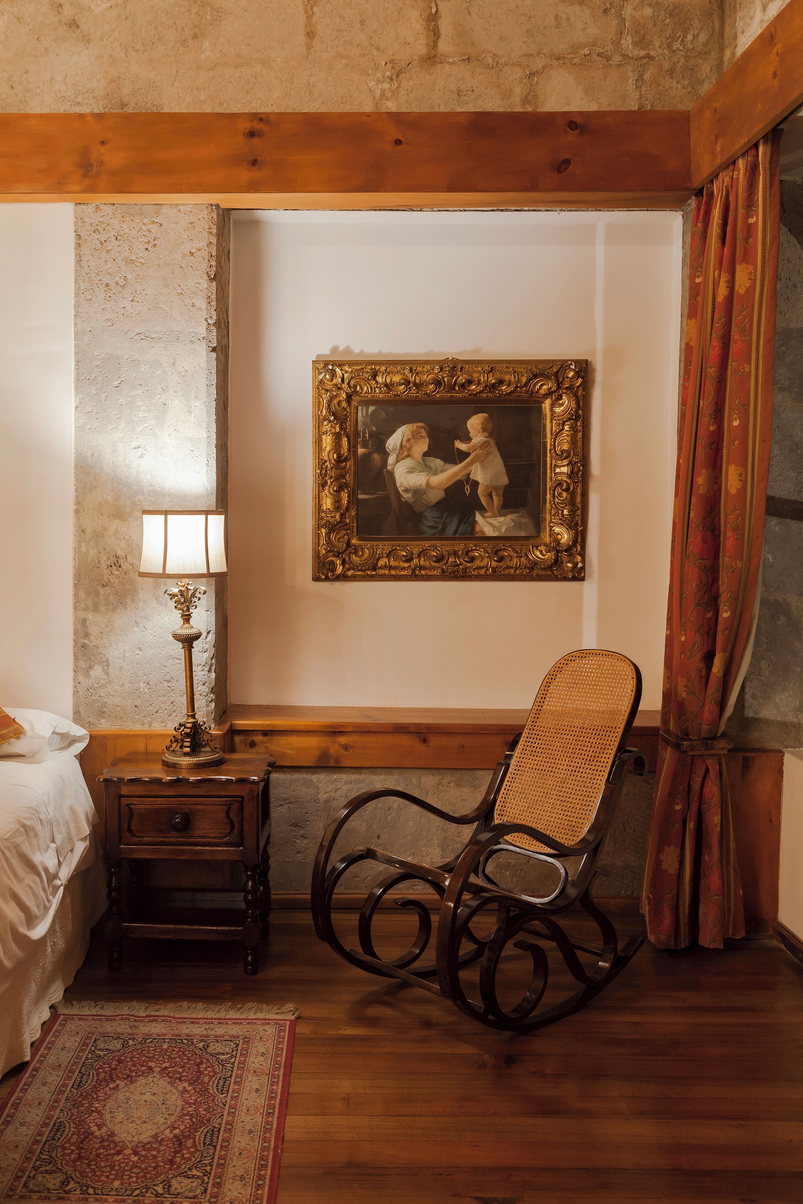 A room with a wooden rocking chair, a small wooden nightstand with a lamp, a framed picture on the wall, and a bed with white bedding.