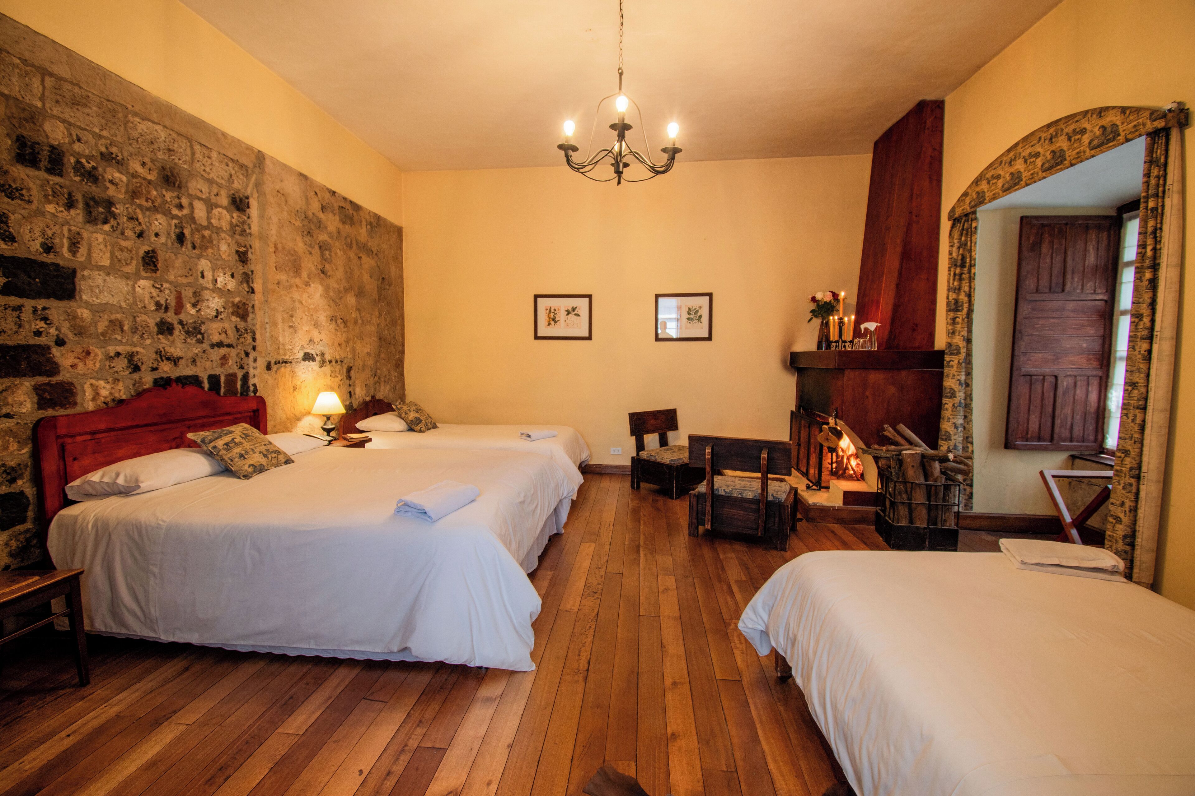 Una habitación con dos camas, piso de madera, pared de piedra, candelabro y espejo con puerta de madera.