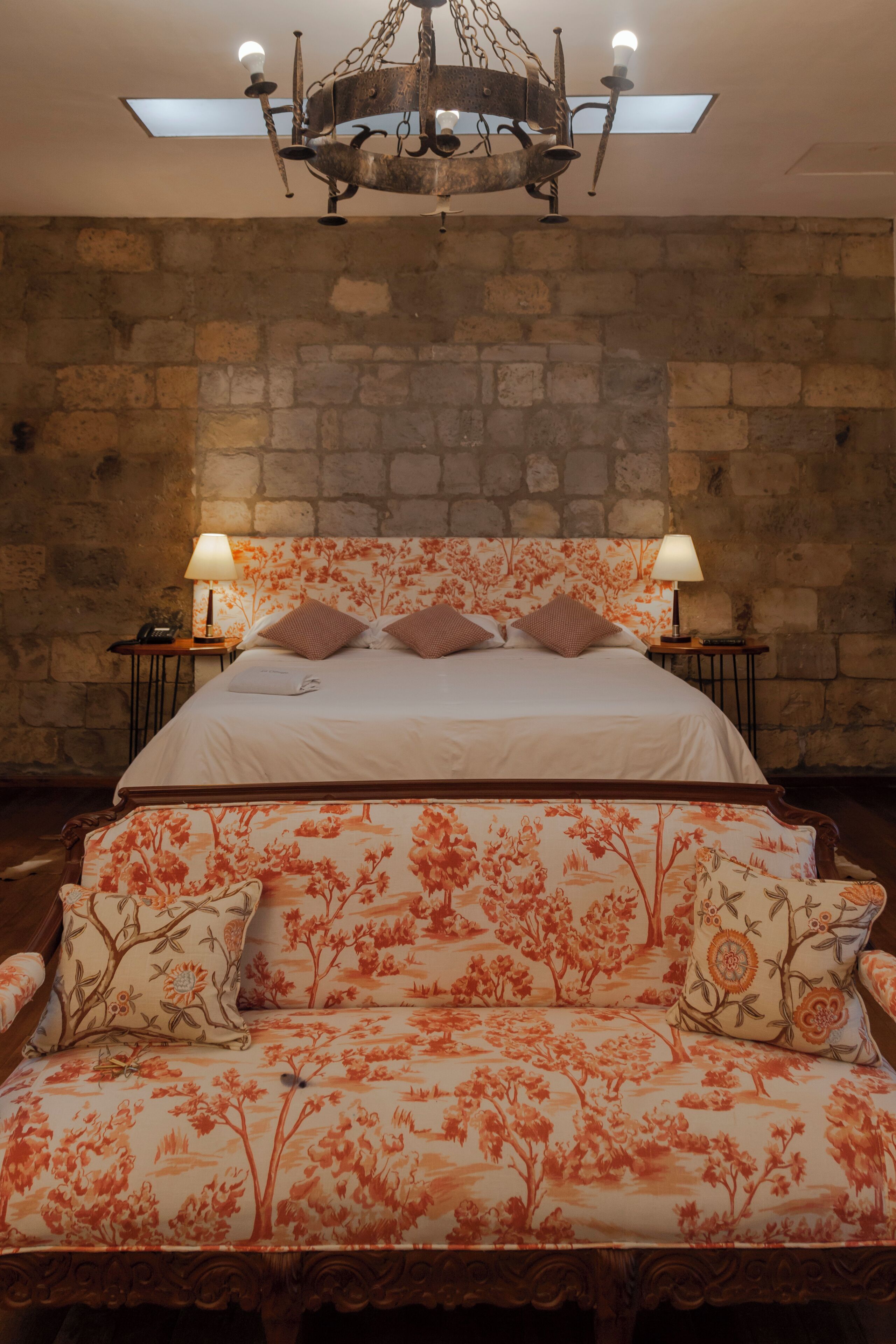 A bedroom with a stone wall, a bed with floral bedding, two bedside lamps, and a chandelier.