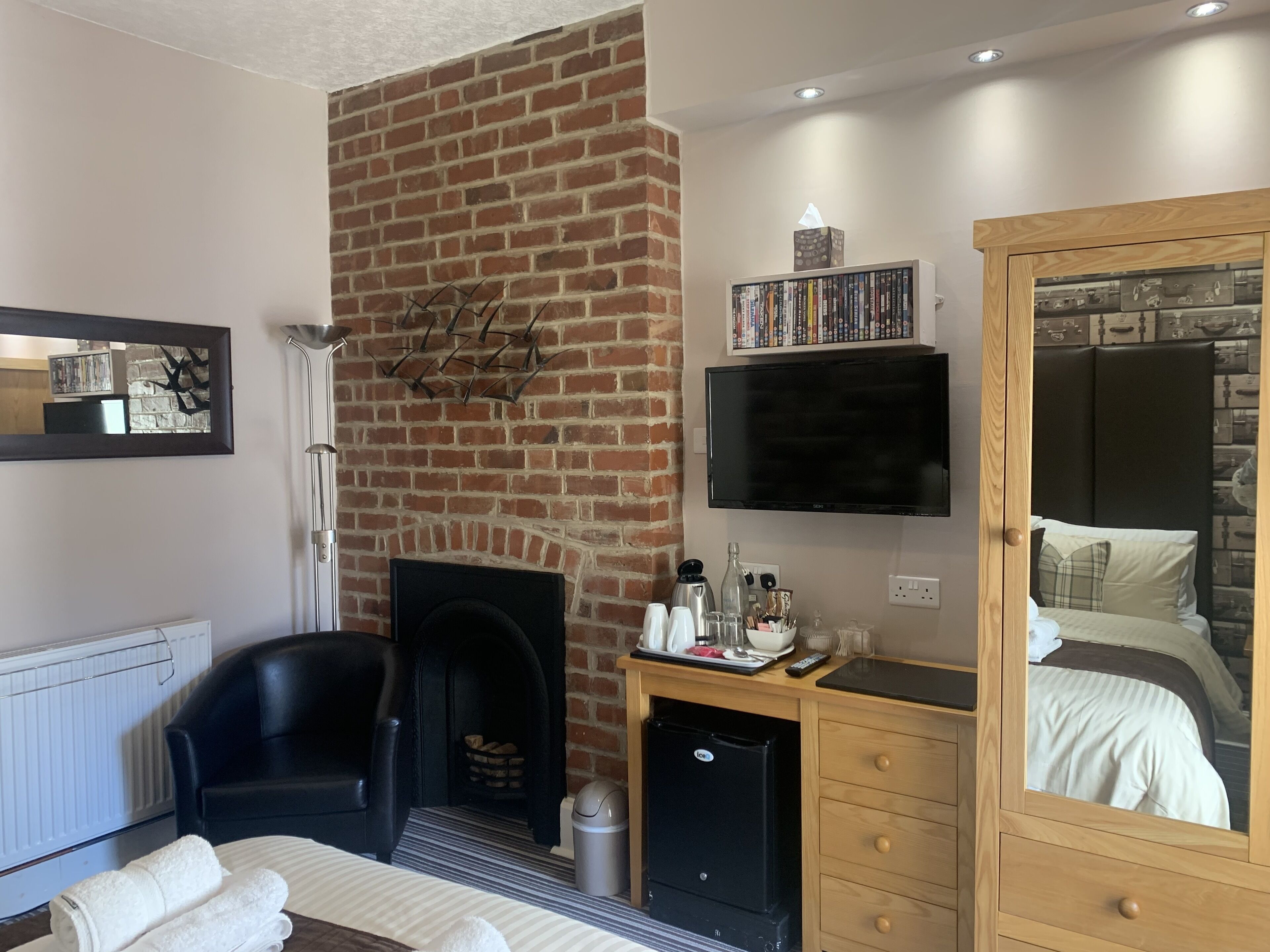 A modern bedroom with a brick fireplace, a TV mounted above it, a wardrobe with a mirror, and a small desk with a lamp.