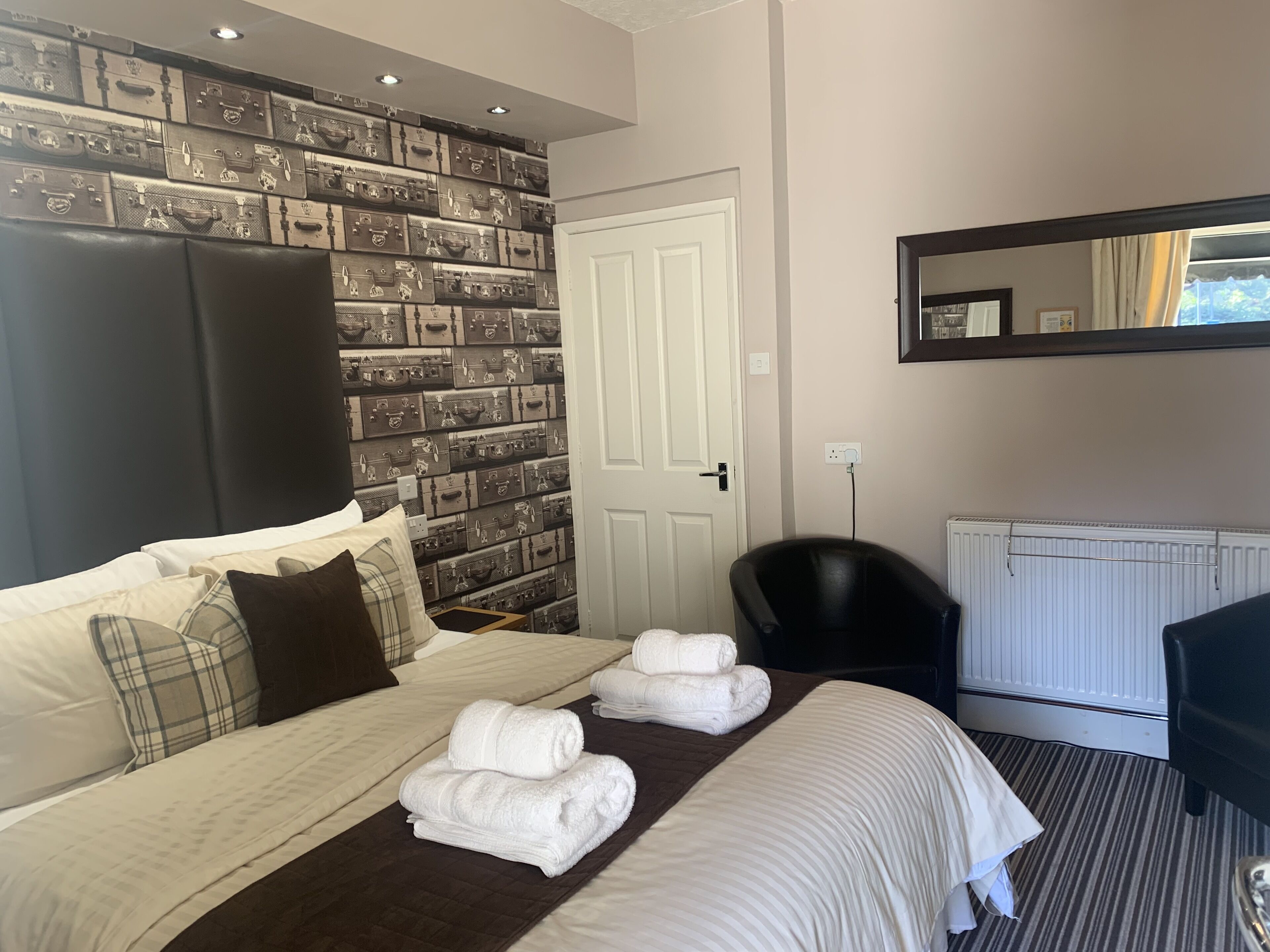 A hotel room with a bed, a wall of suitcases, a black chair, and a radiator.