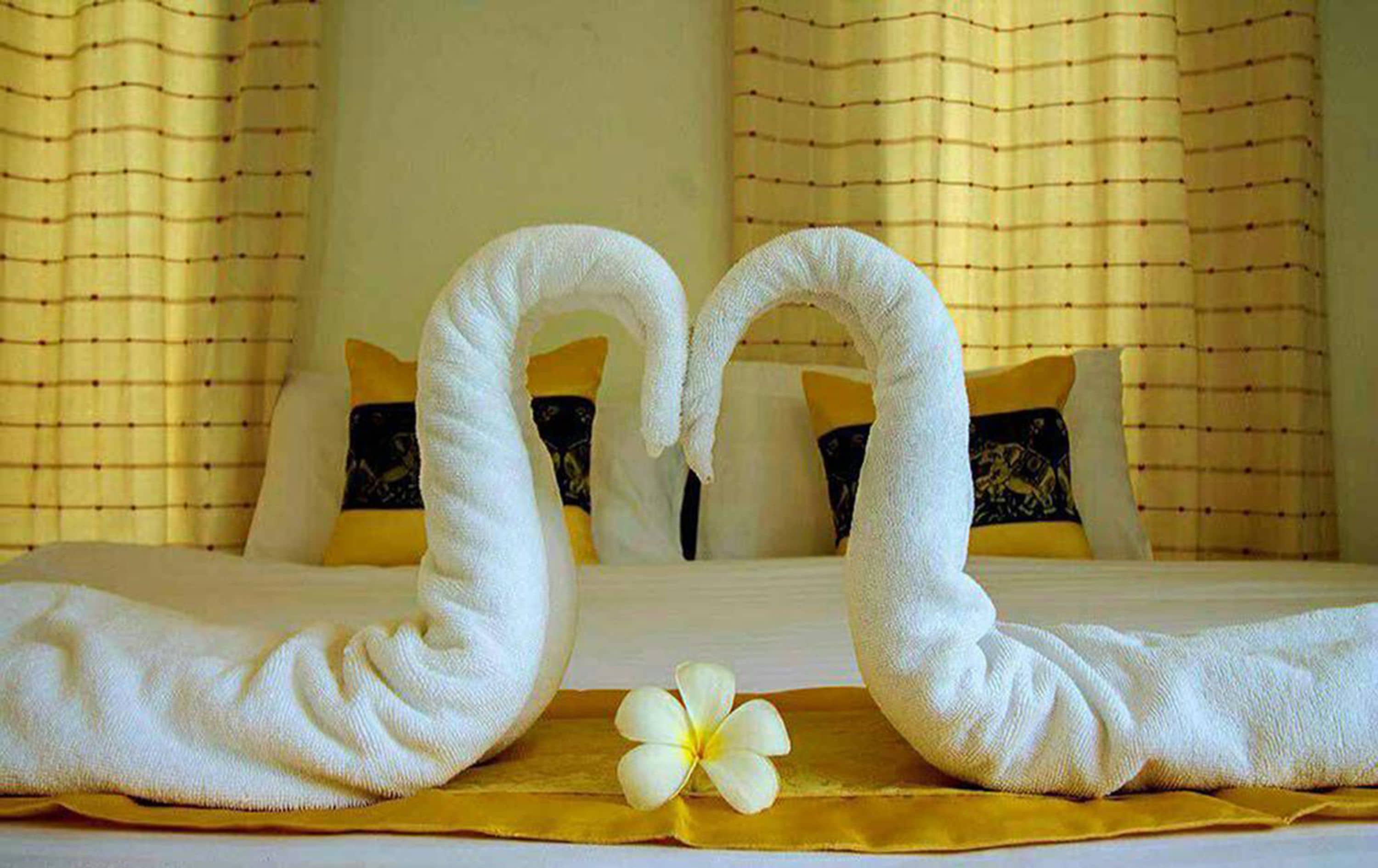 A hotel room with a bed, two towels shaped like swans, and a single white flower on a yellow bedspread.
