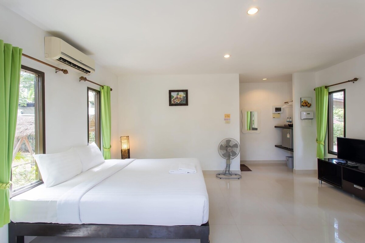 A hotel room with a bed, air conditioning unit, a fan, a TV, and a small table with a lamp.