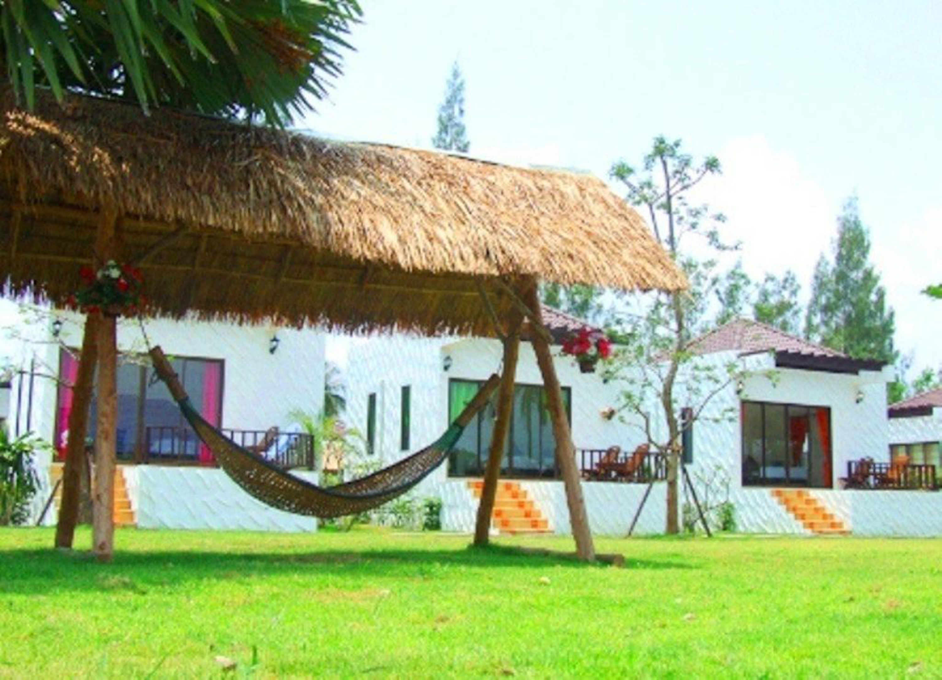A thatched-roof structure with a hammock, white buildings with balconies, and a grassy area.