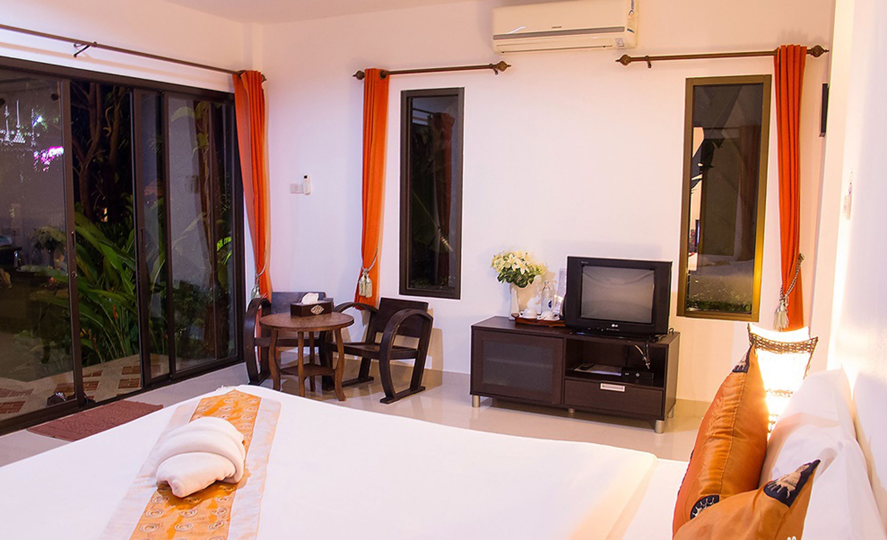 A hotel room with a bed, a TV, a small table with chairs, and a balcony with orange curtains.