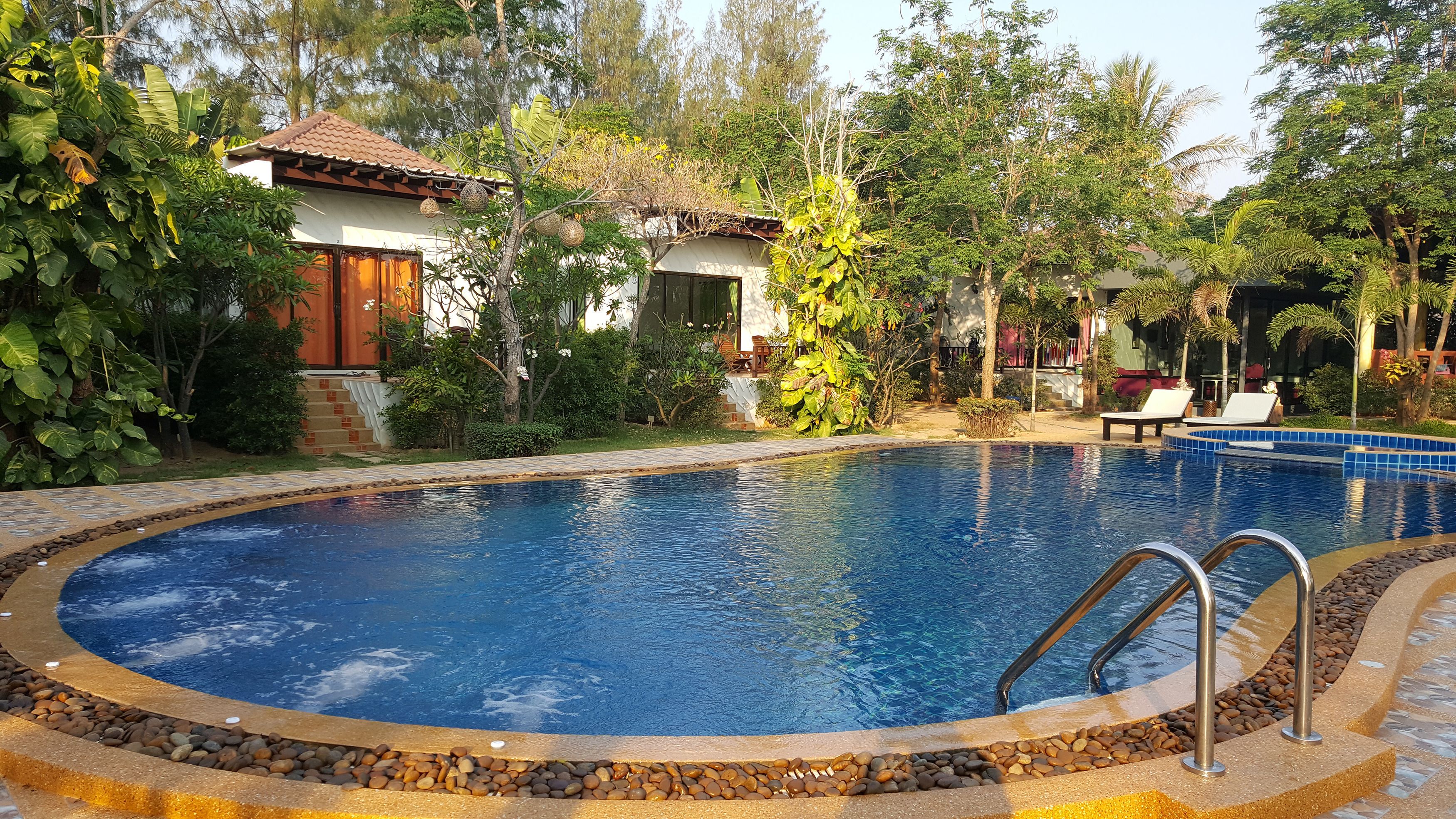 A swimming pool with a stone border, surrounded by lush greenery and a house with a tiled roof.