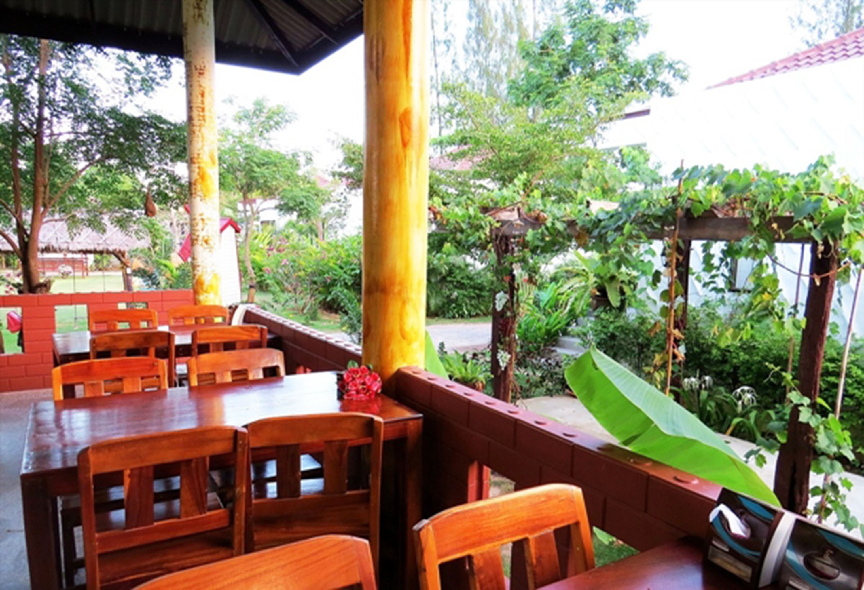 An outdoor dining area with wooden tables and chairs, surrounded by greenery and a pergola.