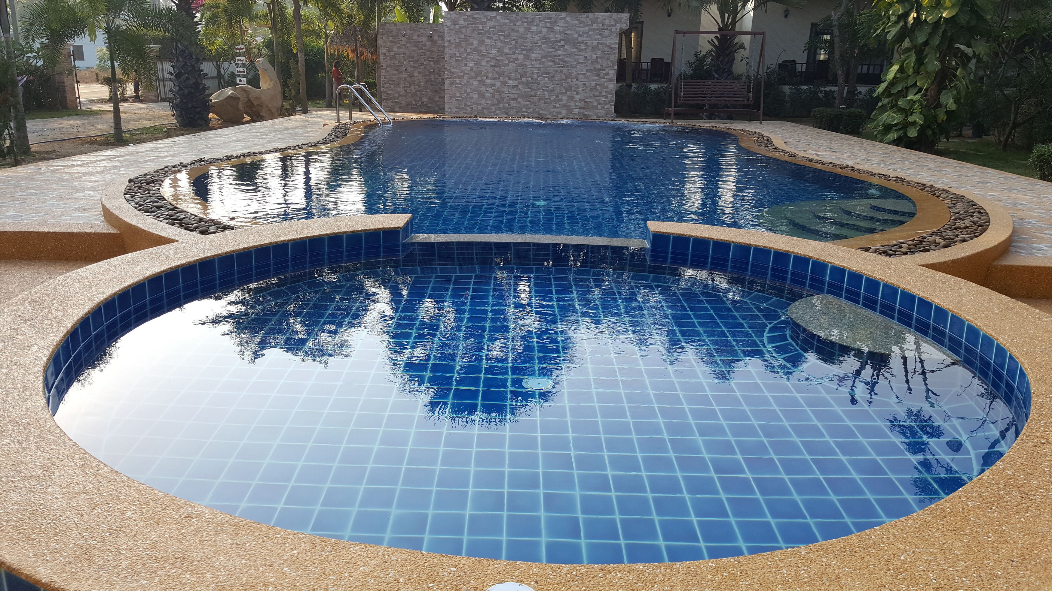 A circular pool with blue tiles and a pebble border, surrounded by a stone wall and palm trees.