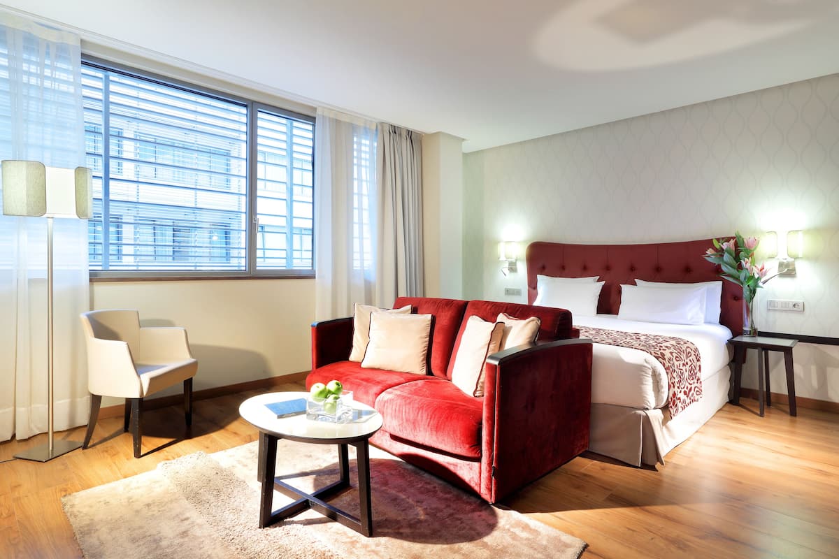 Habitación de hotel con sofá rojo, cama con ropa de cama blanca, mesa de centro redonda con un jarrón y ventana con persianas.