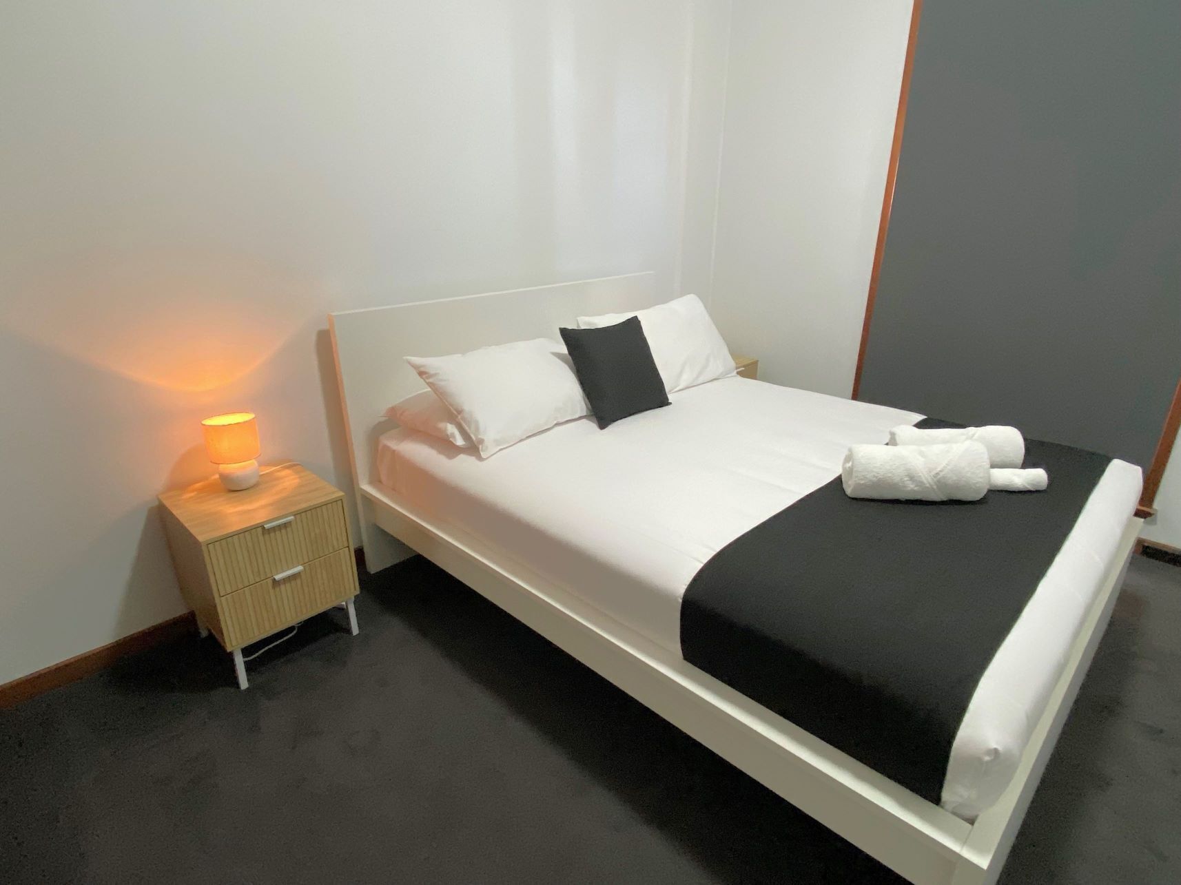 A neatly made bed with white and black bedding, a bedside lamp, and a small wooden nightstand with drawers.