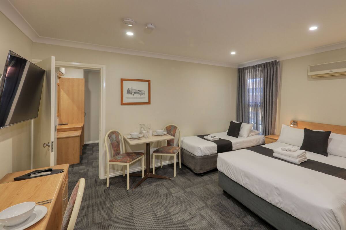 A hotel room with two beds, a desk with chairs, a TV, and an air conditioning unit.