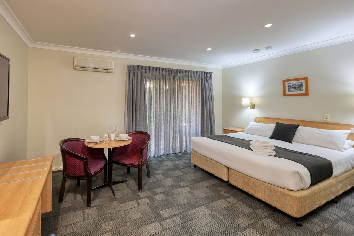 A hotel room with a bed, a desk, two chairs, a table with a bowl and glasses, and a window with curtains.