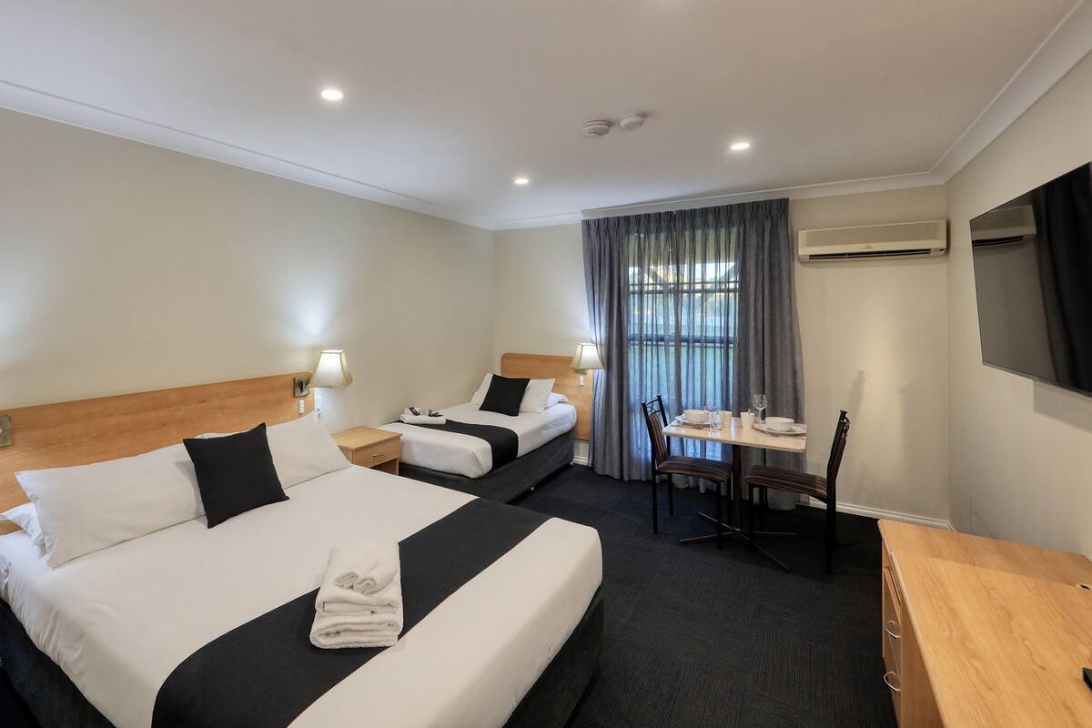 A hotel room with two beds, a desk, chairs, a TV, and an air conditioning unit.