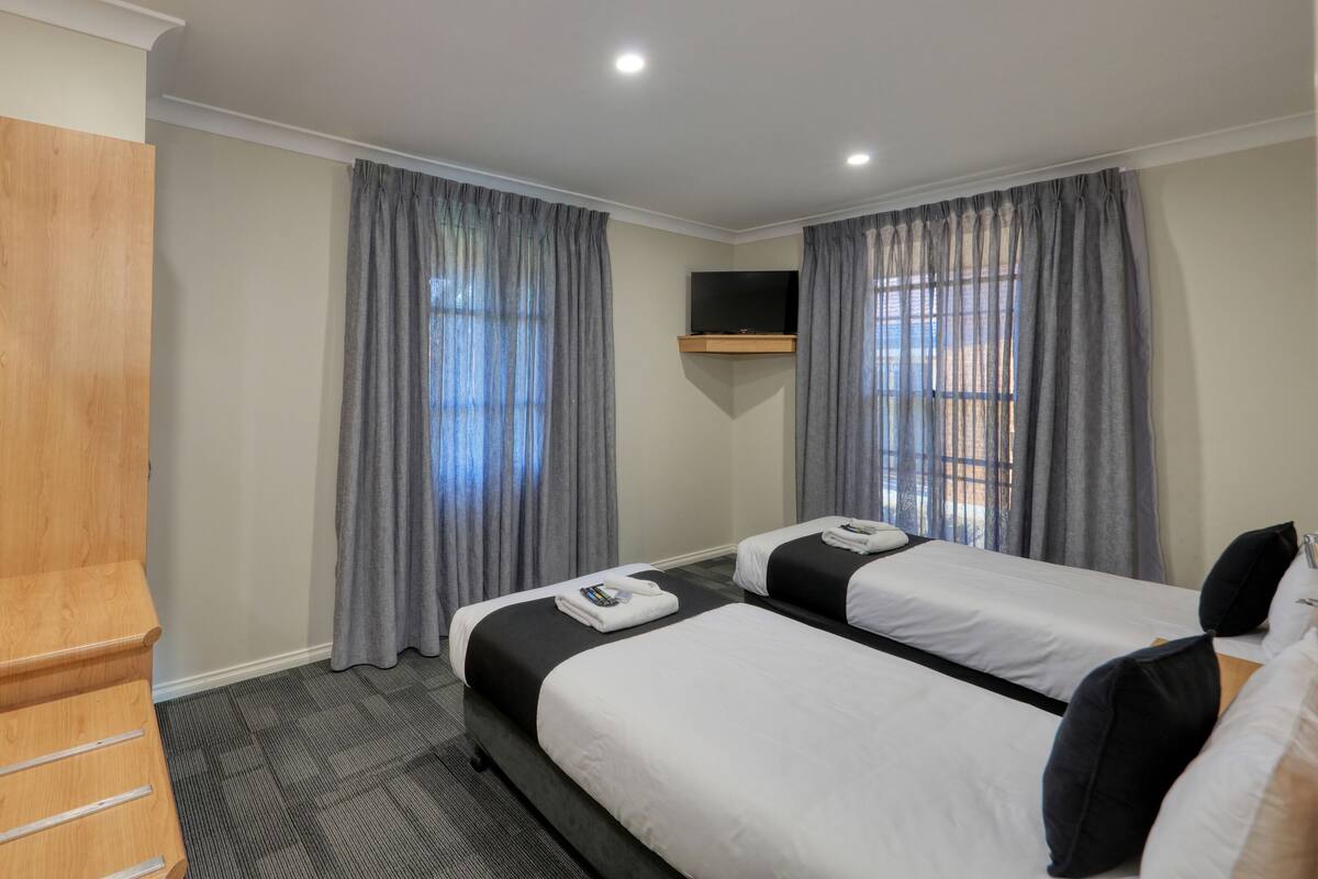 A hotel room with two beds, grey curtains, a wooden bench, and a mounted TV.