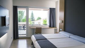 Ein Hotelzimmer mit zwei Betten, einem Schreibtisch, einem Fernseher und einem Fenster mit Blick auf Bäume und Gebäude.