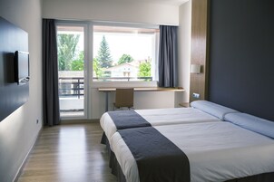 Quarto de hotel com duas camas, uma escrivaninha, televisão e janela com vista para árvores e edifícios.