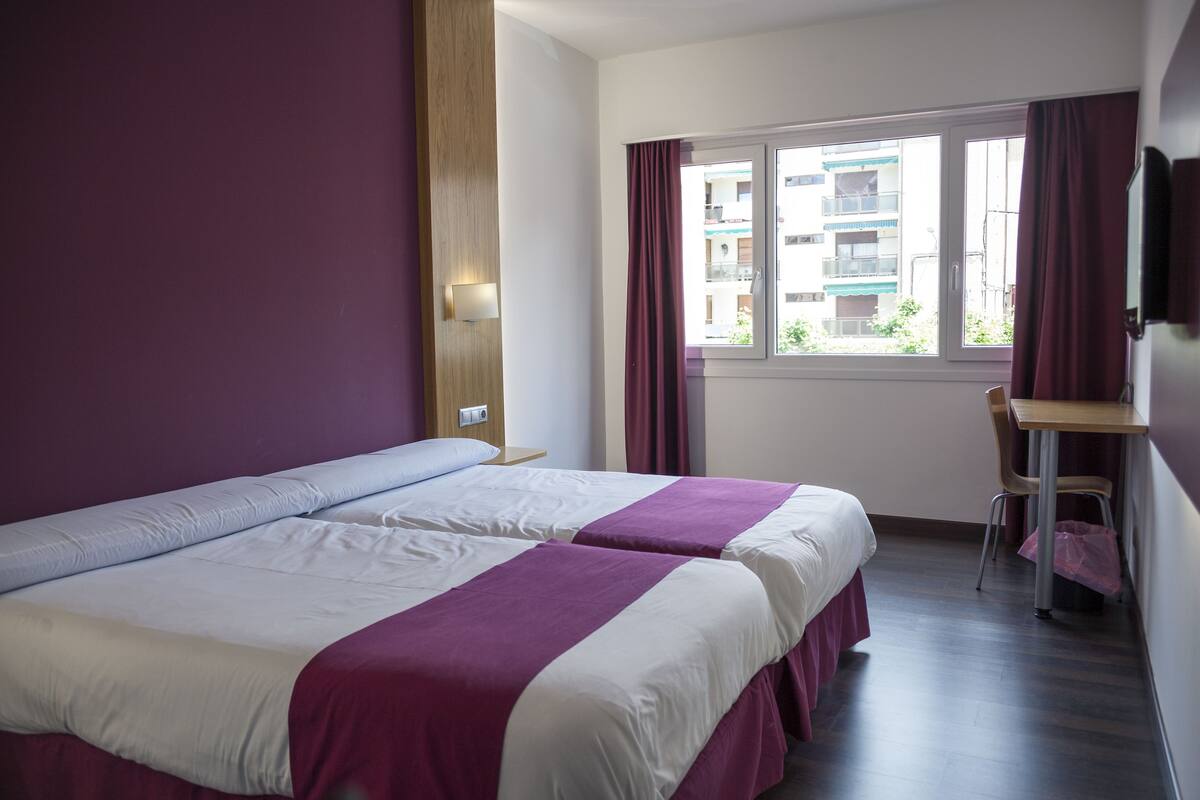 A hotel room with two beds, a desk, a chair, and a window with curtains.