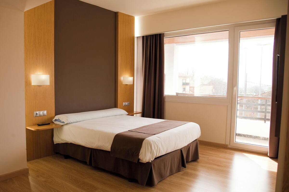A hotel room with a large bed, wooden panel walls, a window with curtains, and a balcony view.