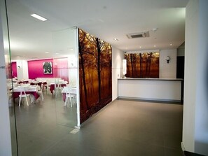A modern restaurant with white tables and chairs, a reception desk, and a forest-themed mural on the wall.