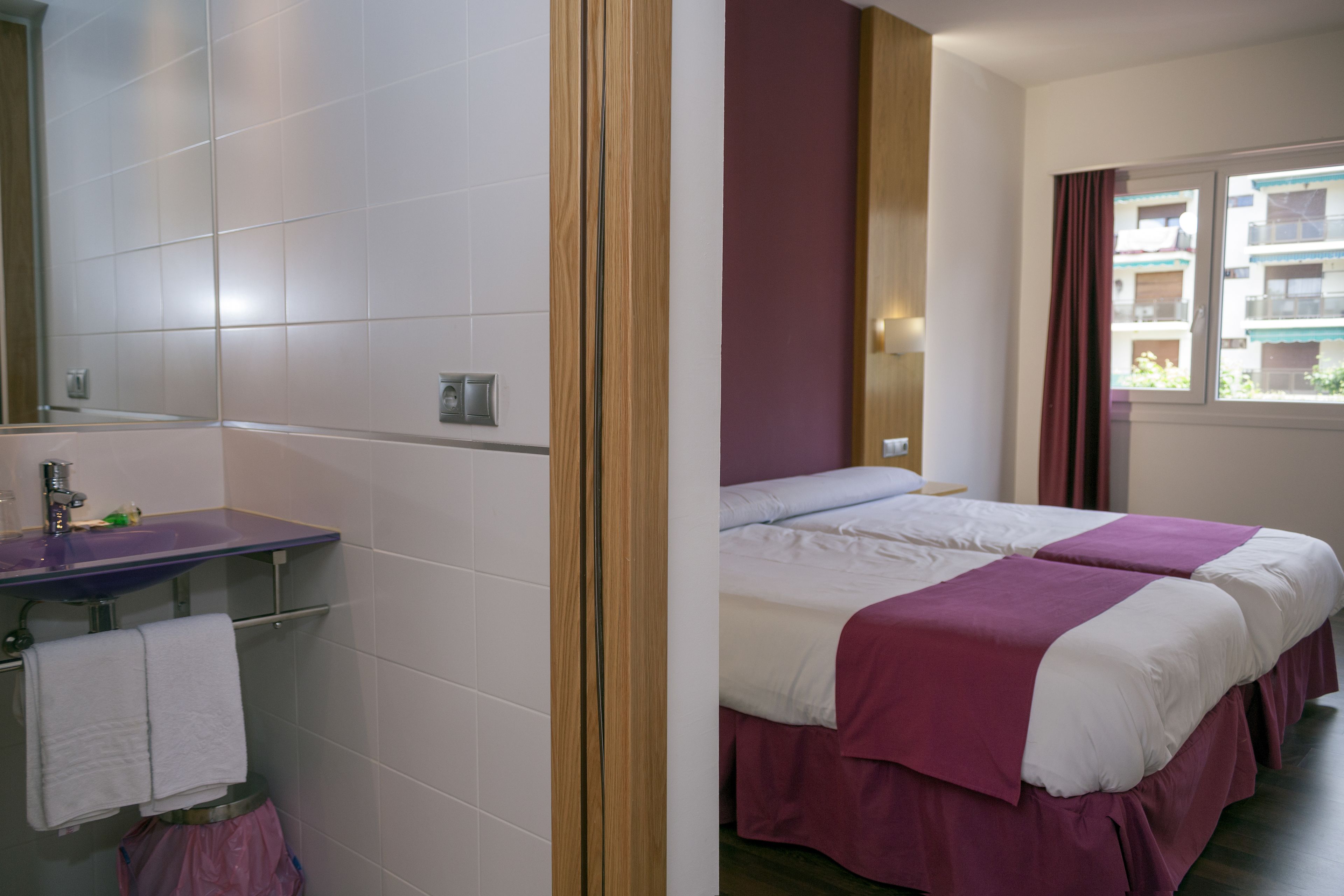 A hotel room with a bed, a bathroom with a sink, and a window with a view of buildings.