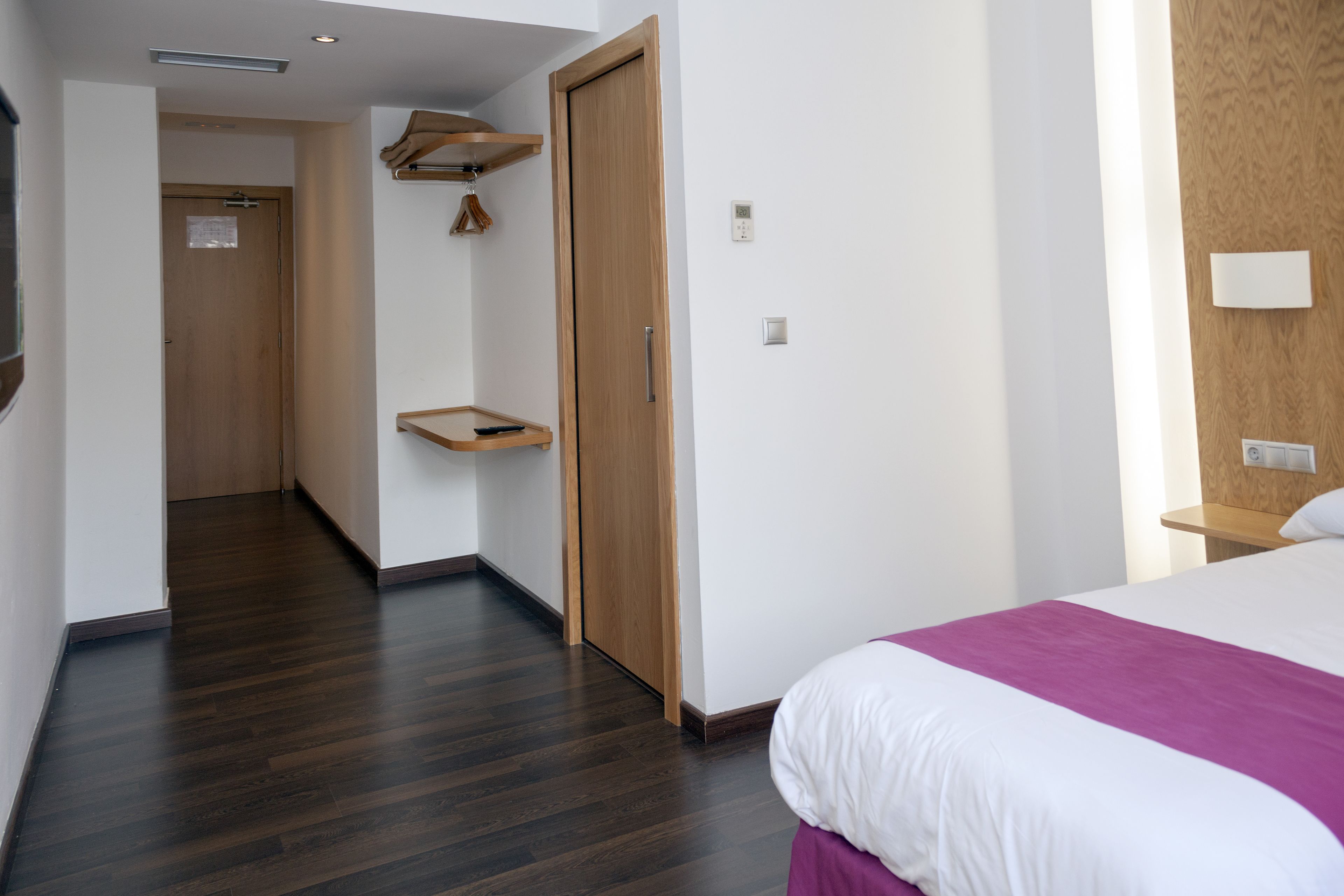 Quarto de hotel com piso de madeira, cama com roupa de cama branca e roxa, guarda-roupa de madeira e uma pequena prateleira de madeira.