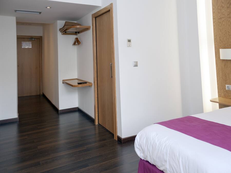 Quarto de hotel com piso de madeira, cama com roupa de cama branca e roxa, guarda-roupa de madeira e uma pequena prateleira de madeira.