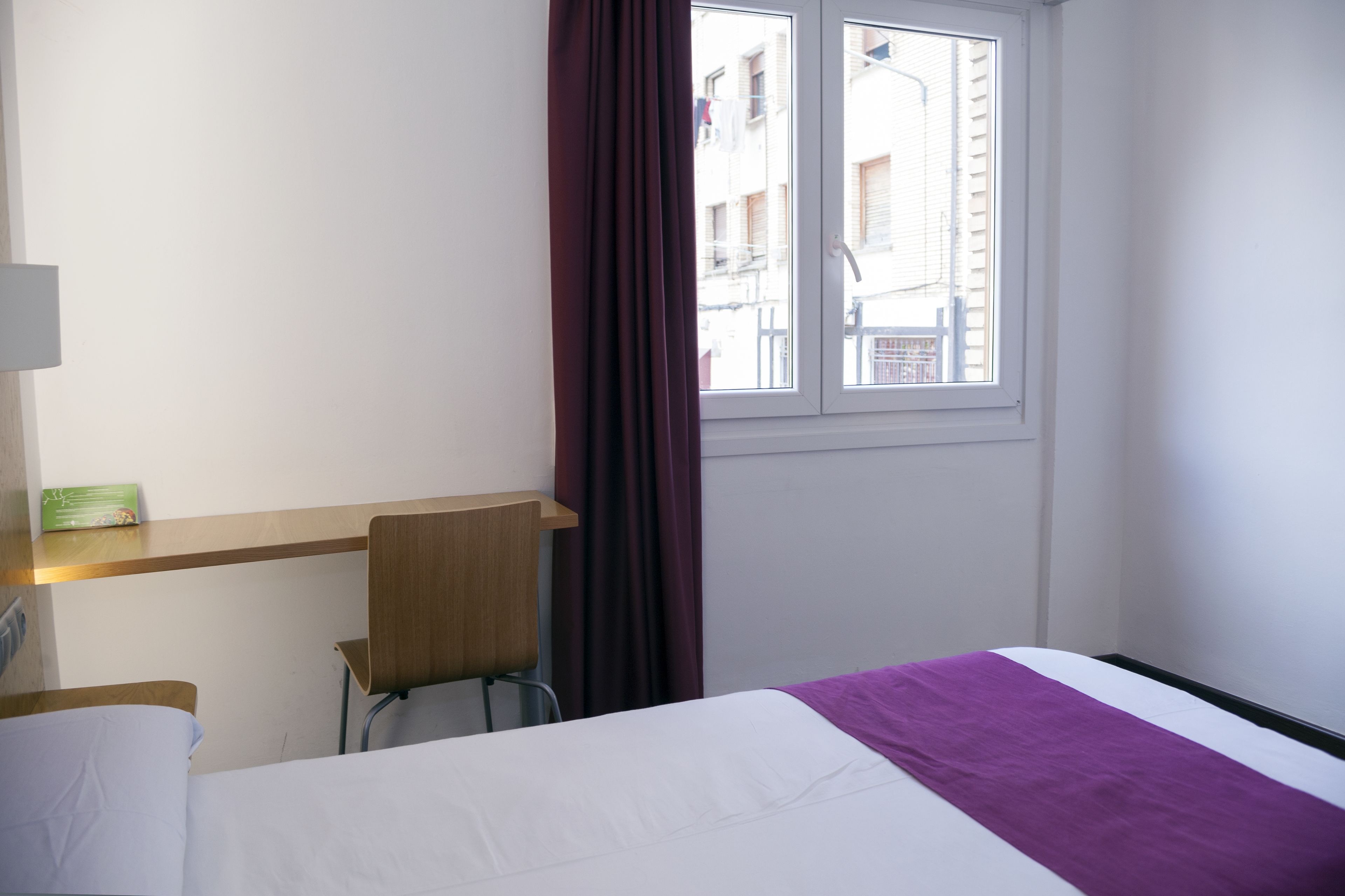 Habitación de hotel con cama, escritorio, silla, ventana con cortinas y vista a edificios.