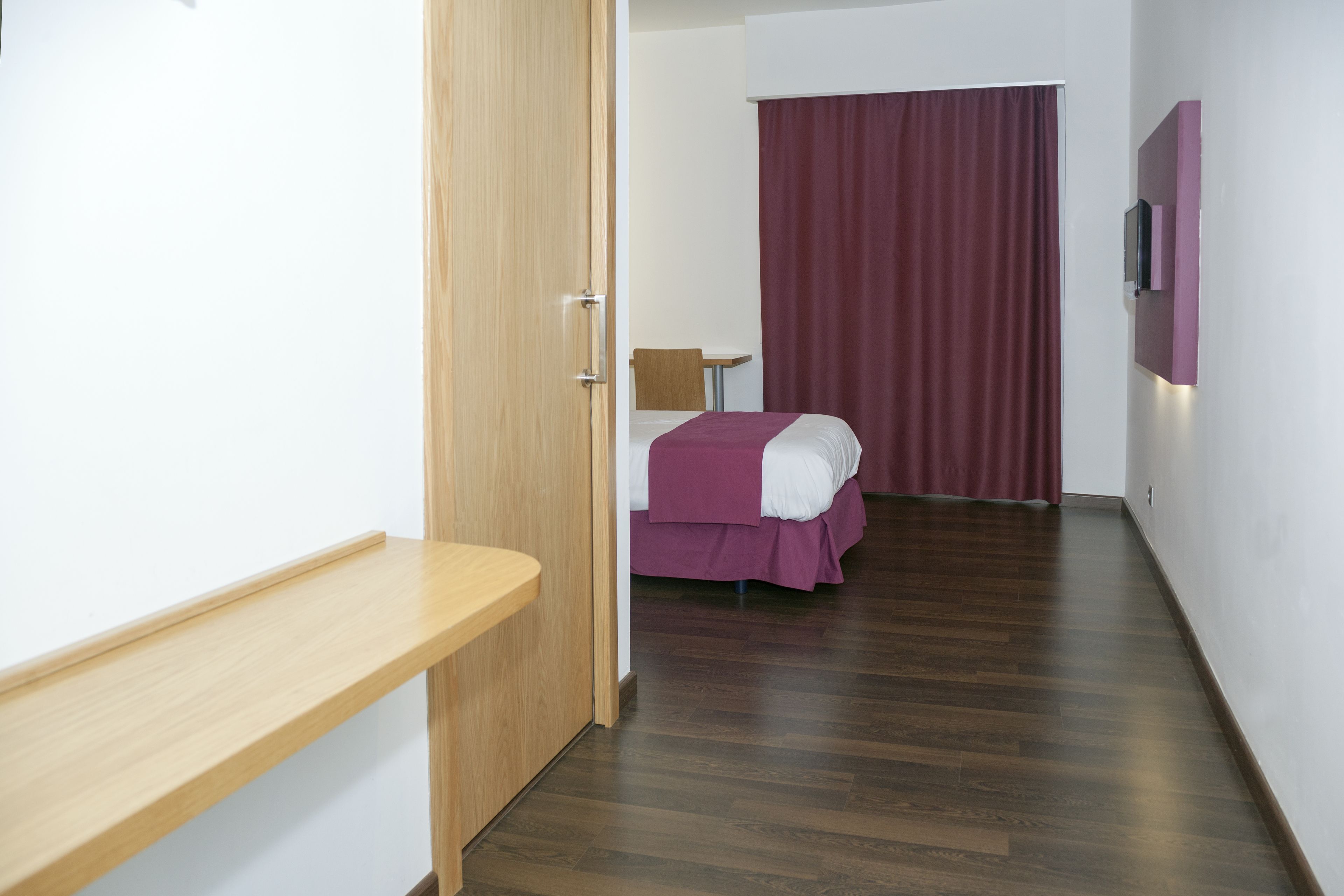 A hotel room with a wooden desk, a bed with a purple bedspread, a small table, and a TV mounted on the wall.