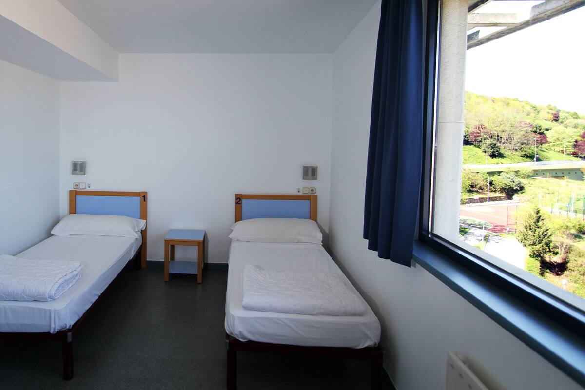 A room with two single beds, a small table, and a window with a view of greenery.