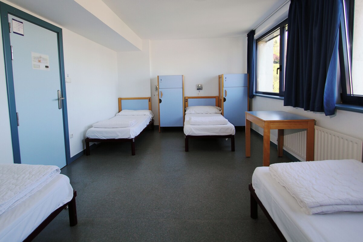 Una habitación con cuatro camas individuales, una mesita, una ventana con cortinas, una puerta y un radiador.