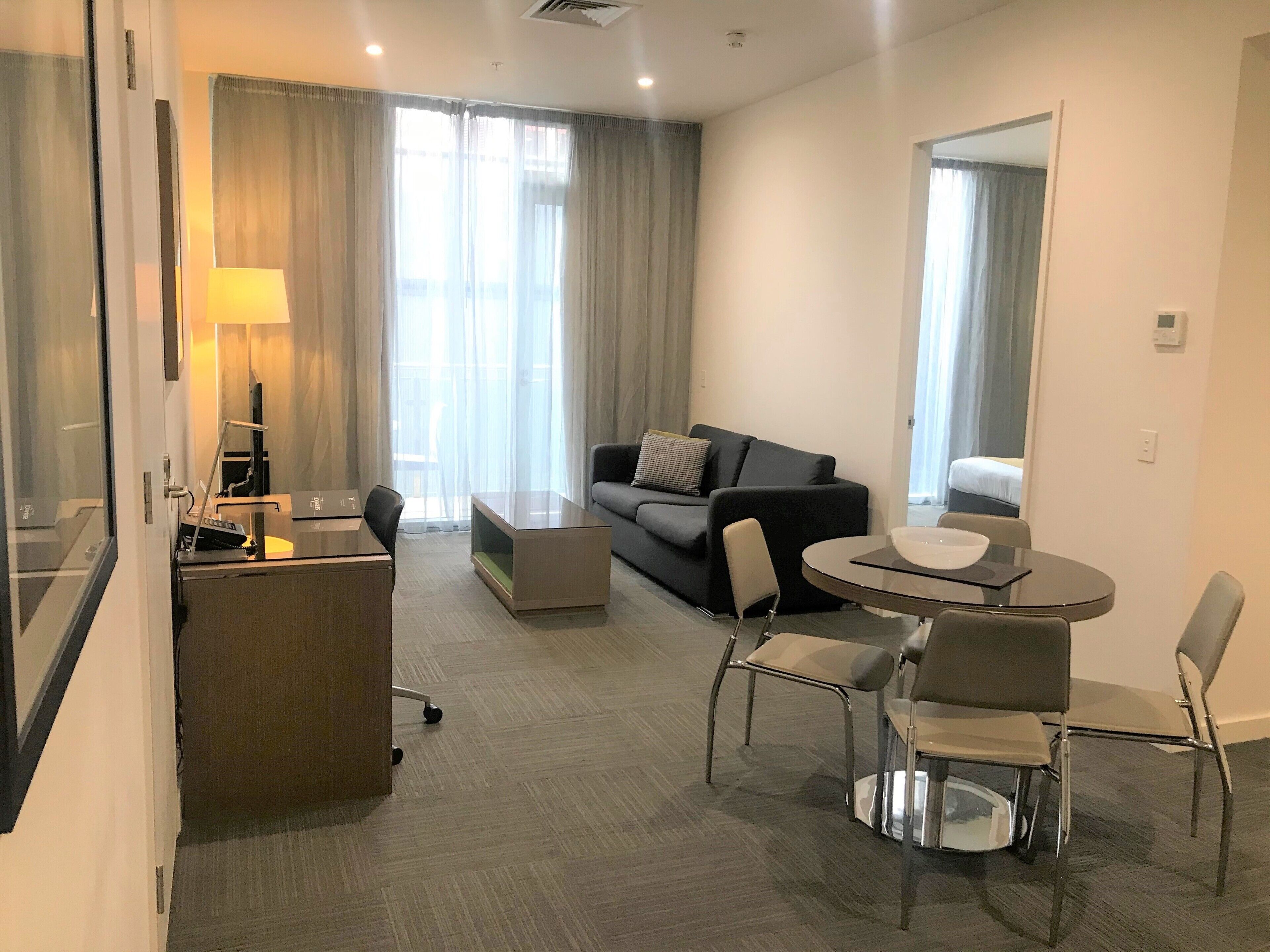A modern hotel room with a sofa, a dining table with chairs, a desk with a lamp, and large windows with curtains.