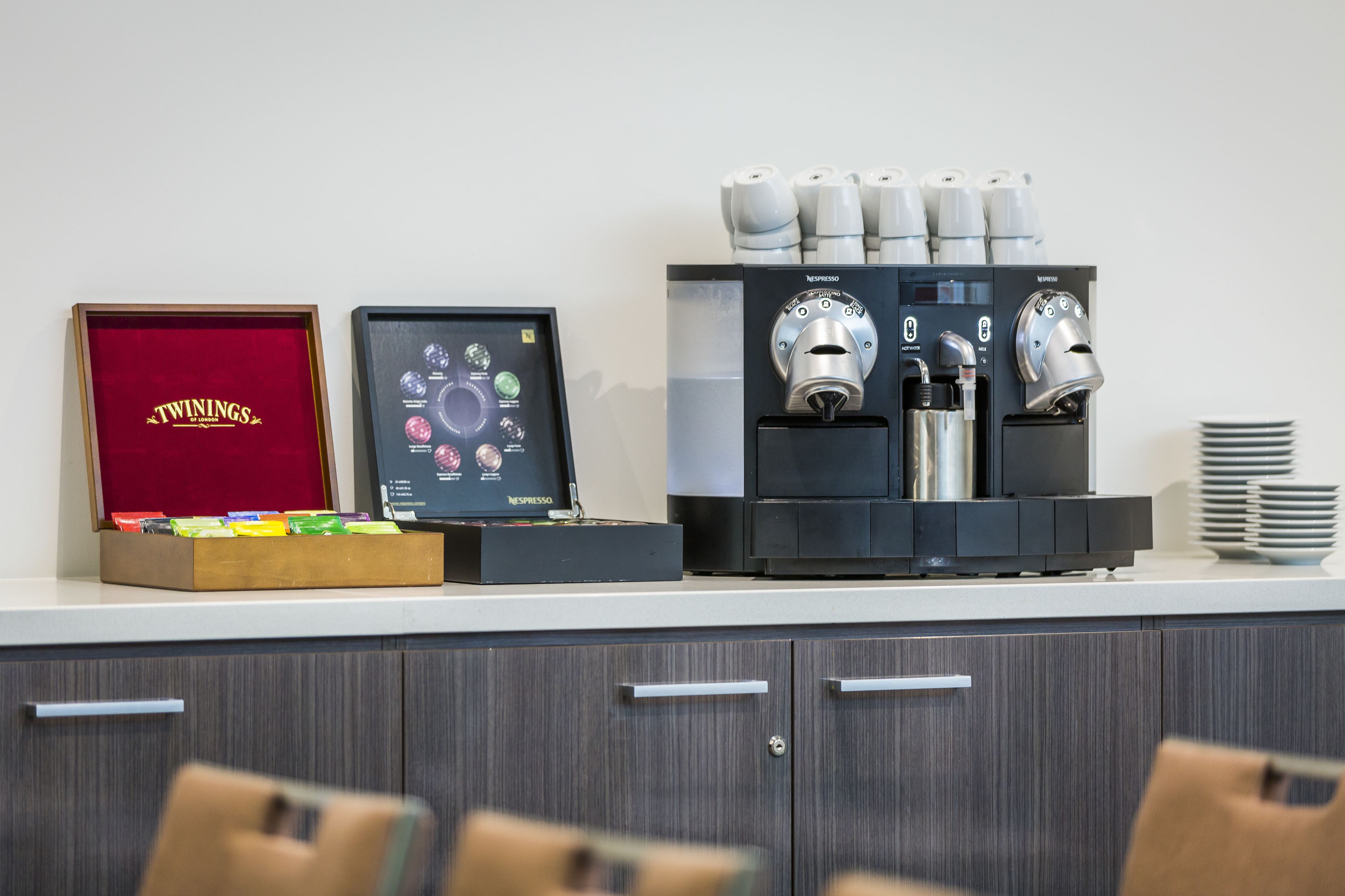 A coffee machine with multiple dispensers and a display screen, a Twinings tea box, and a stack of plates on a counter.