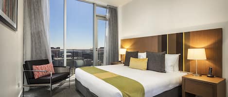 Habitación de hotel con una cama grande, una silla, una mesita de noche y vistas a la ciudad desde la ventana.
