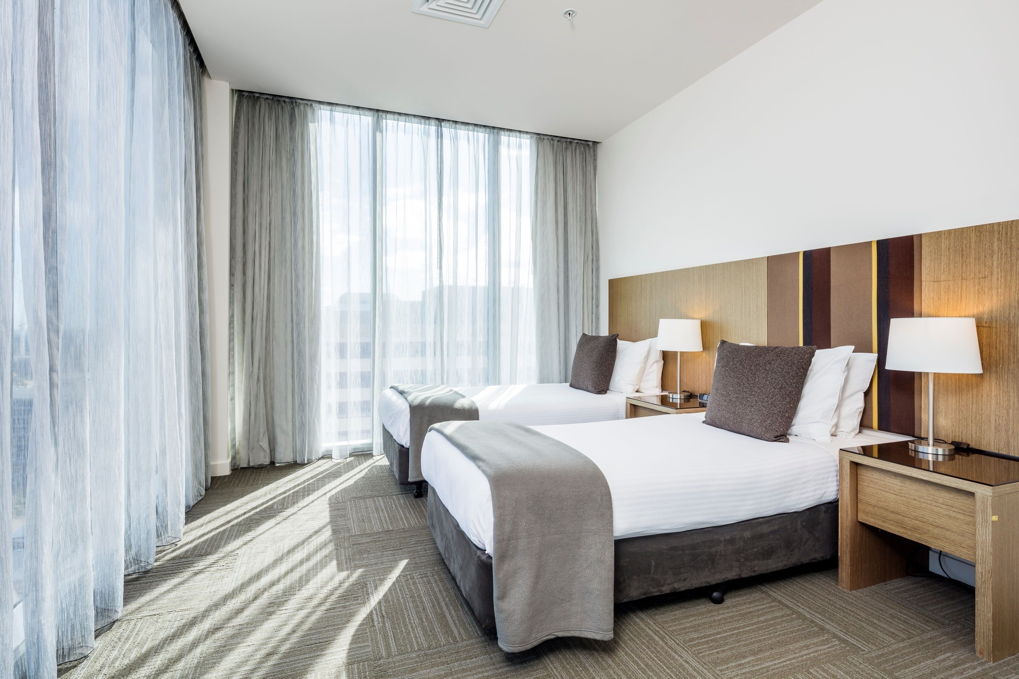 A hotel room with two beds, a large window with sheer curtains, a wooden headboard, and bedside lamps.