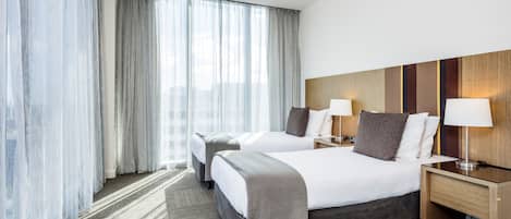 Une chambre d’hôtel avec deux lits, une grande fenêtre avec des rideaux légers, une tête de lit en bois et des lampes de chevet.