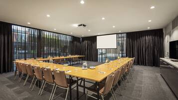 Un salón de conferencias con una larga mesa, sillas, una pantalla de proyector y un amplio ventanal con cortinas.