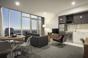 A modern hotel room with a sofa, dining table, chairs, a TV, kitchen area, and large windows offering a city view. - Quest on Franklin (Adelaide)