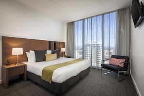 A hotel room with a large bed, a nightstand with a lamp, a chair with a patterned cushion, and a TV mounted on the wall. - Quest on Franklin (Adelaide)