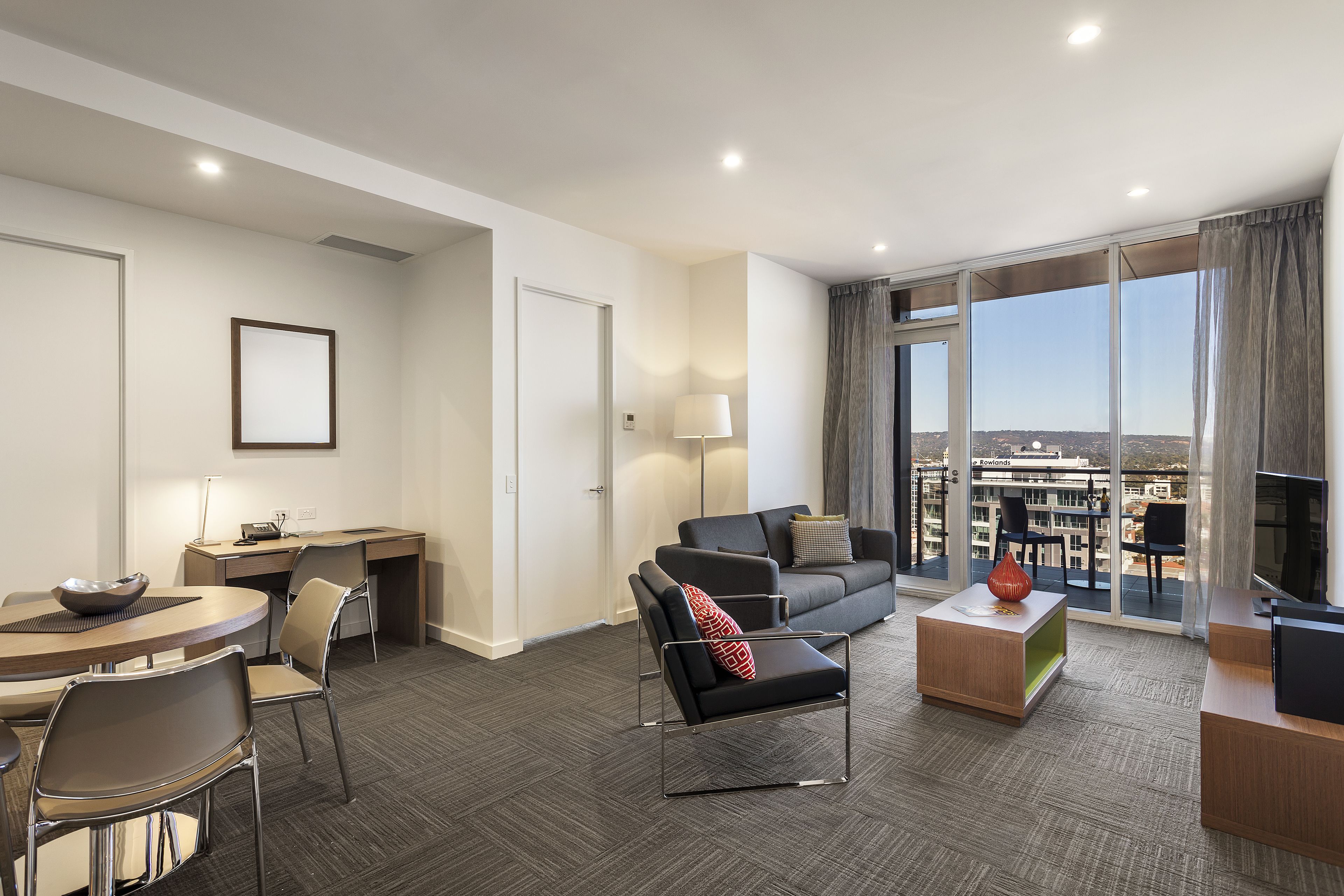 A modern hotel room with a dining area, a sofa, a TV, and a balcony with a city view.
