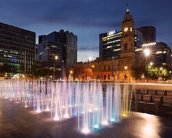 A city square with a historic building featuring a clock tower, modern high-rise buildings, and a fountain with colorful lights.