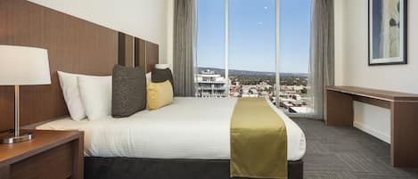 Habitación de hotel con una cama grande, un escritorio y vistas a un paisaje urbano a través de amplios ventanales.