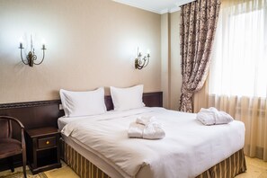 Un letto rifatto con lenzuola bianche, un comodino in legno con un cassetto e una sedia.