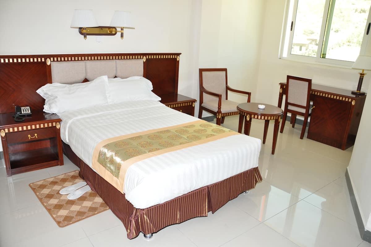 A hotel room with a large bed, a bedside table with a lamp, a chair, a small table, and a window with a view.