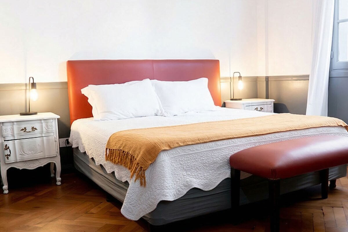 Ein ordentlich bezogenes Bett mit rotem Kopfteil, weißen Kissen und einer beigen Tagesdecke. Zwei Nachttische mit Lampen sowie eine Holzbank am Fußende des Bettes.