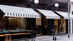 A cafe with striped awnings, a wooden table, and stools outside.