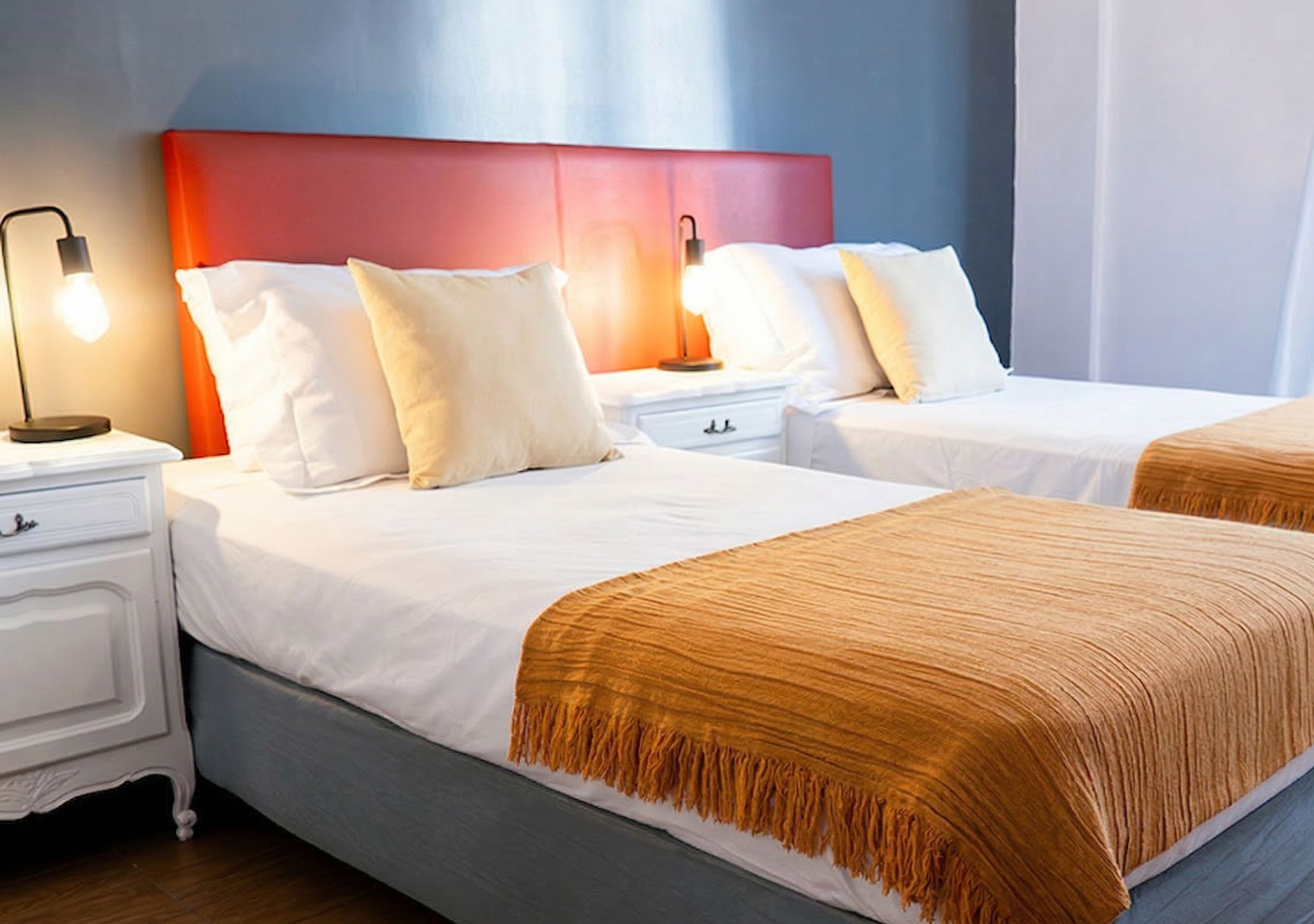 A hotel room with two beds, white bedding, a red headboard, bedside lamps, and a wooden floor.