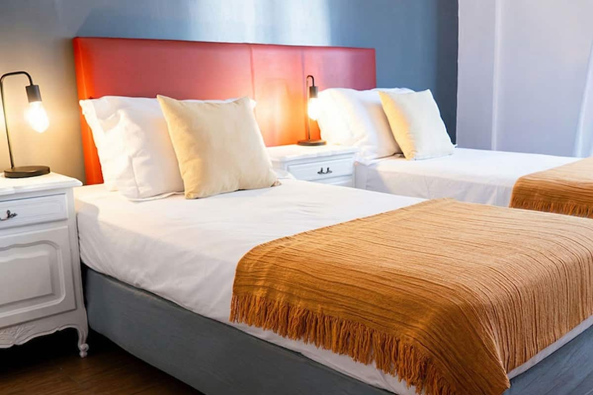 Habitación de hotel con dos camas, ropa de cama blanca, cabecero rojo, lámparas de noche y suelo de madera.