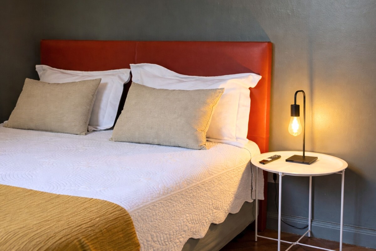 Cama bem arrumada com cabeceira vermelha, roupa de cama branca e uma pequena mesa de cabeceira com abajur.
